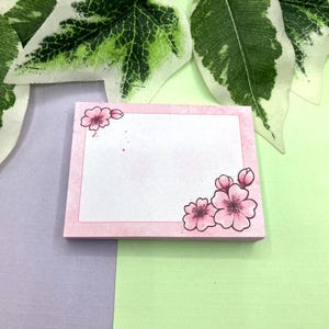 Peut inclure: Blocs de notes adhésifs roses avec un centre blanc et une bordure florale. La bordure présente des fleurs de cerisier roses avec des contours noirs.