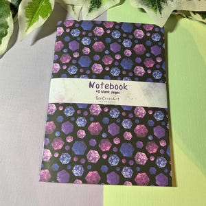 Puede incluir: Un cuaderno con fondo negro cubierto por un patrón de dados morados, rosas y azules. El cuaderno tiene una banda blanca con la palabra "Notebook" y el texto "40 blank pages" impresos.