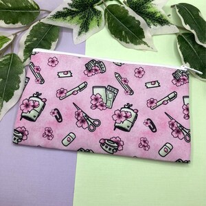 Könnte beinhalten: Rosa Canvas-Federmäppchen mit einem weißen Reißverschluss. Das Federmäppchen ist mit einem sich wiederholenden Muster aus Kirschblüten, Stiften, Scheren und anderen Schreibwaren verziert.