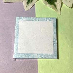 Könnte beinhalten: Quadratischer Notizblock mit hellblauem Rand mit Schneeflockenmustern. Die Mitte des Notizblocks ist weiß mit einem dezenten Schneeflockenmuster. Der Notizblock liegt auf einer lila und grünen Oberfläche.