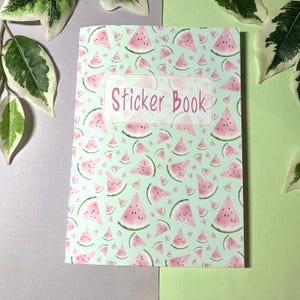 Puede incluir: Un cuaderno de pegatinas con un fondo verde claro y un patrón de rodajas de sandía rosa. Las palabras "Sticker Book" están en rosa sobre una etiqueta rectangular blanca.