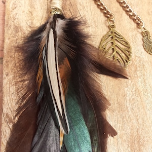 以下が含まれることがあります： 木製の表面からぶら下がっている、ビーズの長いチェーン。茶色と緑の羽根のチャームと、金の葉のチャームが2つ付いている。