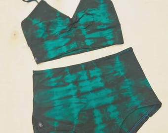 May include: A two-piece set of underwear with a tie-dye pattern in shades of green and black. The top is a bralette with thin straps and a knot detail. The high-waisted bottoms have a blue stitch detail. A tag is attached.