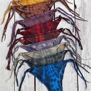 May include: A stack of seven colorful tie-dye bikini bottoms with adjustable ties. The bottoms are made of a soft, stretchy fabric and feature a variety of tie-dye patterns.