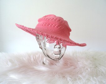 Könnte beinhalten: Ein pinkfarbener Sonnenhut mit breiter Krempe und Häkeldesign. Der Hut wird auf einem transparenten Glaskopf vor weißem Hintergrund präsentiert. Der Rand hat einen gewellten Rand und die Krone ein strukturiertes Muster.