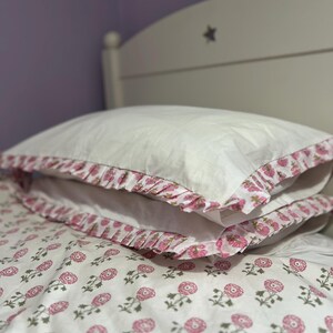 Puede incluir: Juego de cama blanco con un estampado floral rosa. Las fundas de almohada tienen un borde con volantes con el mismo diseño floral. El marco de la cama es blanco con un recorte de estrella.