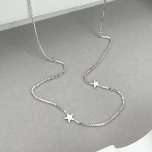 May include: A delicate silver necklace featuring a box chain design. The necklace has two small star pendants. The jewelry is displayed on a gray surface.
