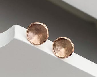 May include: A pair of rose gold stud earrings with a hammered, textured finish. The earrings are round and have a slightly concave shape.