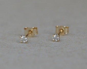 May include: A pair of small, gold stud earrings, each featuring a single, clear, round-cut gemstone. The earrings are set on a gold post with a butterfly backing, displayed against a neutral background.