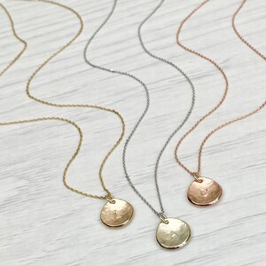 K&ouml;nnte beinhalten: Drei goldene Halsketten mit geh&auml;mmerten Scheibenanh&auml;ngern, jeweils mit einem anderen Anfangsbuchstaben: L, A und C. Die Halsketten liegen auf einem hellen Holzhintergrund.