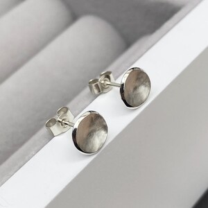 May include: A pair of silver stud earrings with a round, slightly domed shape. The earrings have a brushed finish.