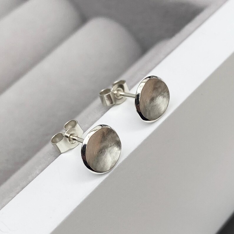May include: A pair of silver stud earrings with a round, slightly domed shape. The earrings have a brushed finish.