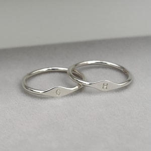May include: Two silver rings with a minimalist design. Each ring features a small, diamond-shaped detail with a single letter engraved on it. One ring has the letter "G", and the other has the letter "H". The rings are set against a light grey background.