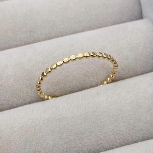 May include: A thin gold ring featuring a continuous band of small, rounded, bead-like spheres. The metal has a polished, reflective finish, creating a delicate and uniform circular design that maintains a consistent texture throughout the entire band.
