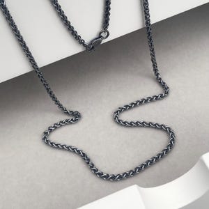 May include: A dark grey wheat chain necklace featuring a woven, textured link design. The metal chain has a polished finish and includes a secure lobster claw clasp mechanism at the top, connecting the two ends of the continuous, flexible metal strand.