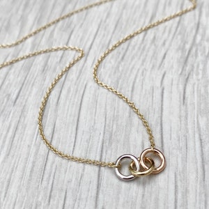 May include: A delicate gold chain necklace with a three-ring pendant. The rings are interlocked and made of different shades of gold, including white, yellow, and rose gold.