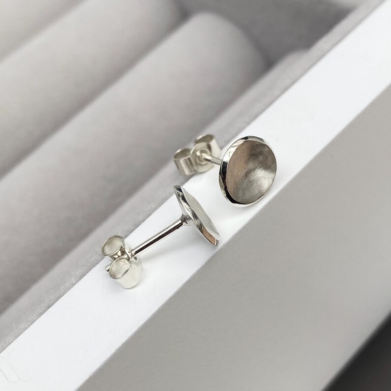 May include: A pair of silver stud earrings with a round, slightly concave design. The earrings are displayed in a white box.