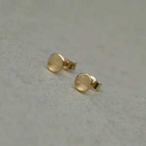 May include: A pair of small, round, gold earrings. The earrings are simple and elegant, with a slightly textured surface.