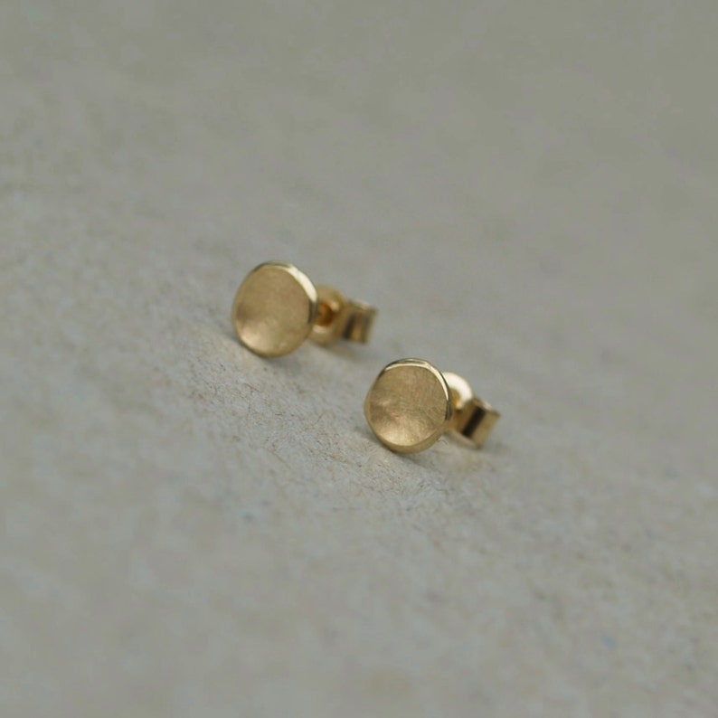 May include: A pair of small, round, gold earrings. The earrings are simple and elegant, with a slightly textured surface.
