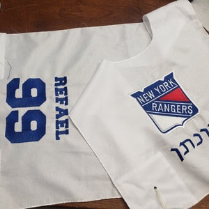 Peut inclure: Tissu blanc avec broderie bleue du numéro 99 et du nom "REFAEL". Un tissu blanc avec un logo bleu et rouge des Rangers de New York et le mot hébreu "לזכרון" brodé en bleu.