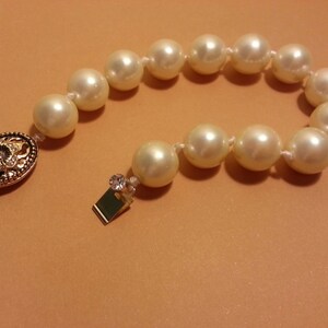 May include: A white pearl bracelet with a gold clasp featuring a floral design. The bracelet is made of faux pearls and has a simple, elegant design.