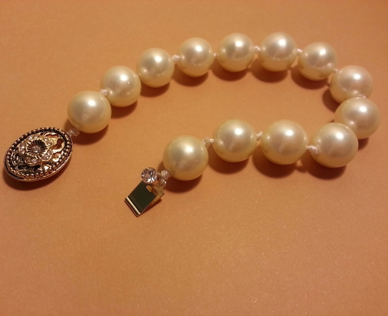 May include: A white pearl bracelet with a gold clasp featuring a floral design. The bracelet is made of faux pearls and has a simple, elegant design.