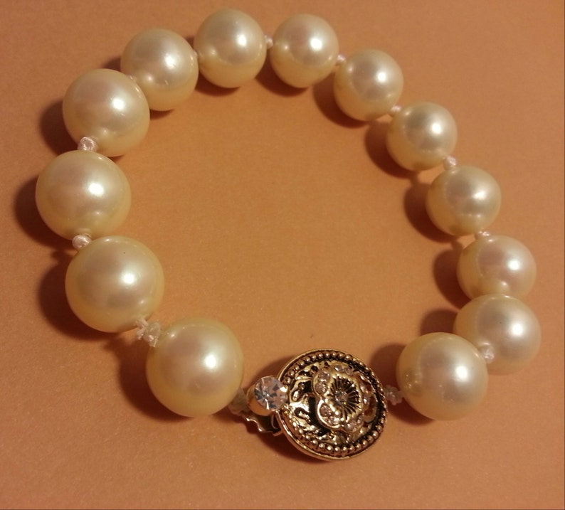 May include: A white pearl bracelet with a gold clasp. The clasp has a decorative design and a small clear rhinestone.