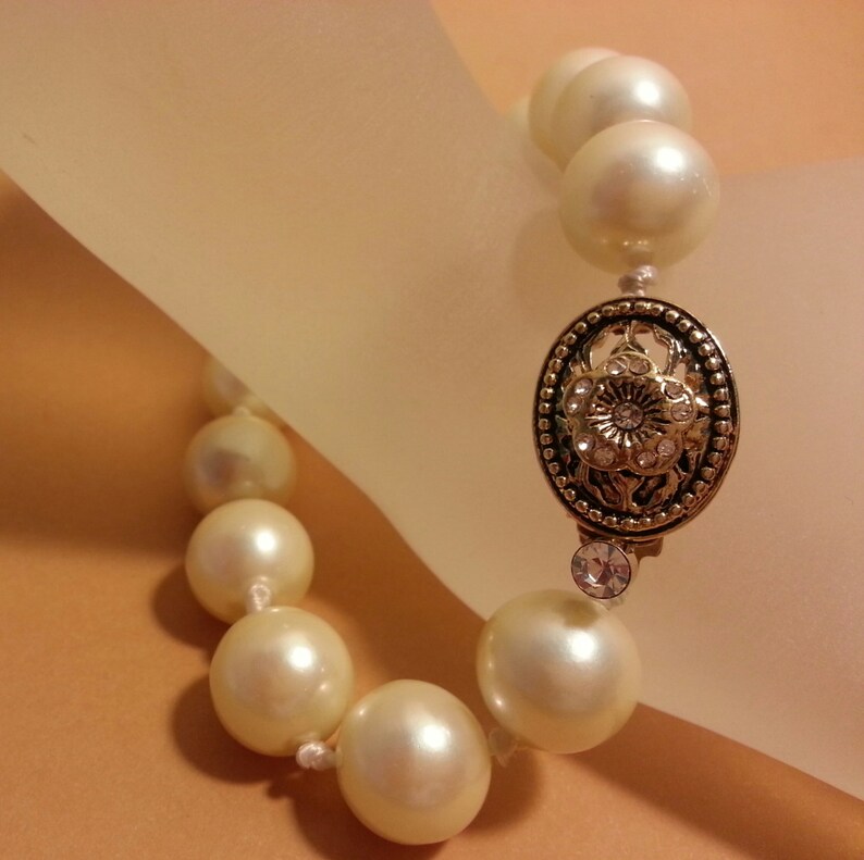 May include: A pearl bracelet with a gold clasp featuring a floral design and a small rhinestone. The pearls are white and the bracelet is on a light background.