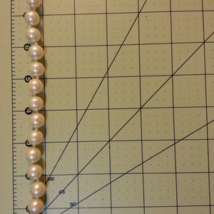 May include: A white pearl necklace with a gold clasp. The necklace is laid out on a gridded surface.