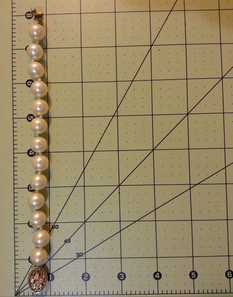 May include: A white pearl necklace with a gold clasp. The necklace is laid out on a gridded surface.