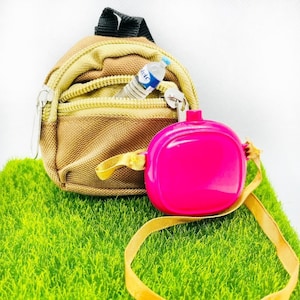 Puede incluir: Una pequeña mochila marrón con un bolsillo con cremallera y una botella de agua azul en su interior. La mochila está unida a un cordón rosa y dorado.