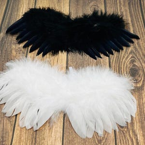 May include: Two sets of decorative feather wings, one black and one white, are displayed against a wooden background. The wings are designed with a layered feather construction, creating a soft and textured appearance. The black wings are positioned above the white wings.
