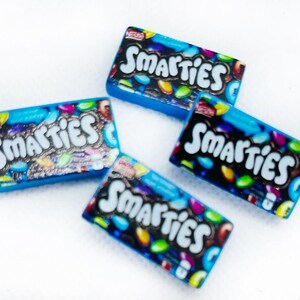 May include: Four miniature boxes of Smarties candy. The boxes are blue with a white background and feature the Smarties logo and colorful candies.