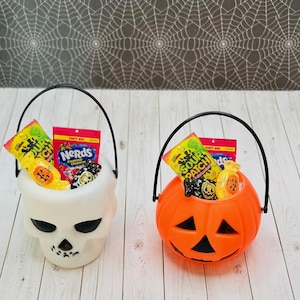 May include: Two Halloween trick-or-treat buckets, one shaped like a skull and the other like a pumpkin, filled with candy. The skull bucket is white and the pumpkin bucket is orange. The candy includes Nerds gummy clusters, Sour Patch Kids, and other assorted candies.