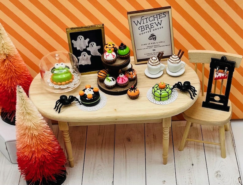 K&ouml;nnte beinhalten: Ein Miniatur-Halloween-Caf&eacute; mit einem Holztisch und St&uuml;hlen. Der Tisch ist mit verschiedenen Halloween-Cupcakes, Kuchen und Kaffee dekoriert. Das Schild &uuml;ber dem Caf&eacute; lautet "Witches Brew Coffee House".