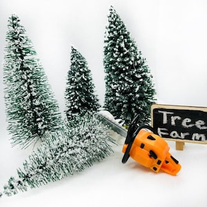 May include: A miniature orange chainsaw with a black handle and a silver blade, resting on a white surface with three small, snow-covered evergreen trees and a black chalkboard sign that reads "Tree Farm".