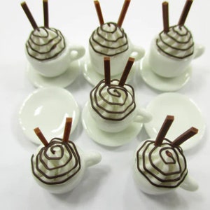 K&ouml;nnte beinhalten: Satz mit sechs Miniatur-Kaffeetassen aus wei&szlig;em Keramik mit Schokoladenwirbel-Schlagsahne und zwei Schokoladenst&auml;bchen obenauf. Jede Tasse steht auf einer wei&szlig;en Untertasse.