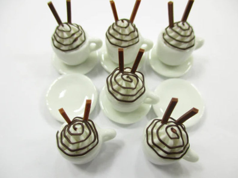 K&ouml;nnte beinhalten: Satz mit sechs Miniatur-Kaffeetassen aus wei&szlig;em Keramik mit Schokoladenwirbel-Schlagsahne und zwei Schokoladenst&auml;bchen obenauf. Jede Tasse steht auf einer wei&szlig;en Untertasse.