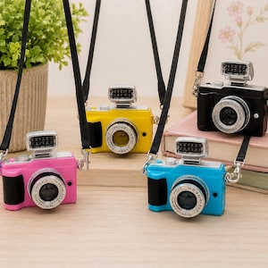 Puede incluir: Cuatro cámaras de juguete en miniatura de color rosa, amarillo, negro y azul, cada una con un objetivo plateado, un flash montado en la parte superior y una correa negra. Los objetivos llevan la inscripción OPTICALS 14-42mm 1:3.5-5.6 y otras marcas.