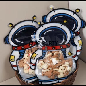 May include: Three clear plastic bags with astronaut designs filled with small, colorful candies. The bags are in a brown wicker basket.