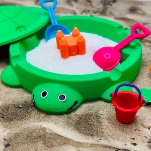 May include: A green turtle-shaped sandbox with a lid, filled with white sand. Includes a pink shovel, blue shovel, orange castle, and a red bucket. The sandbox is on a sandy surface.