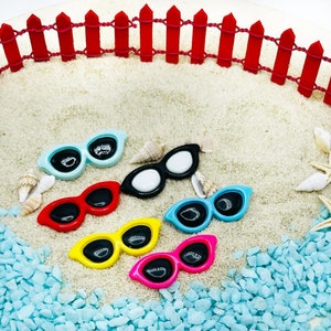 Könnte beinhalten: Sechs Paare farbenfroher Miniatur-Sonnenbrillen auf einer weißen Sandfläche mit blauen und weißen Kieselsteinen. Die Sonnenbrillen sind in verschiedenen Farben, darunter Blau, Schwarz, Rot, Gelb und Pink.