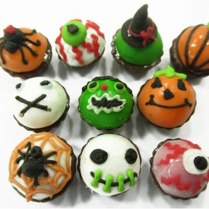 K&ouml;nnte beinhalten: Eine Sammlung von zehn Halloween-Cupcakes, die mit verschiedenen gruseligen Designs verziert sind, darunter eine Spinne, ein K&uuml;rbis, ein Geist und ein Auge. Die Cupcakes sind alle in verschiedenen Farben glasiert, darunter Orange, Gr&uuml;n, Wei&szlig; und Pink.