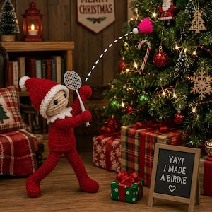 May include: A crocheted elf doll in a red outfit and hat holding a miniature badminton racket, hitting a pink shuttlecock toward a decorated Christmas tree. A small chalkboard sign in the foreground displays the text: YAY! I MADE A BIRDIE.