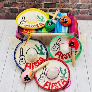 Puede incluir: Cuatro mini sombreros coloridos con la palabra "Fiesta" bordada en ellos, y tres maracas con diseños coloridos. Los sombreros y las maracas están sobre una superficie de madera blanca.