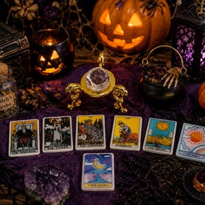 May include: A collection of seven rectangular tarot cards featuring The Emperor, The Tower, The Empress, Strength, The Moon, and The Sun, with a clear faceted crystal sphere on a gold stand, a jar labeled Witch's Herbs, and Halloween decor.