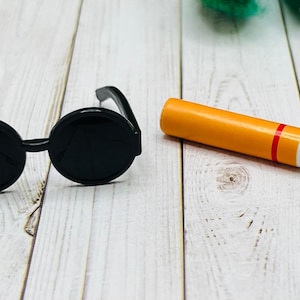 Puede incluir: Gafas de sol redondas negras y un cigarrillo de juguete con una banda roja y blanca y una punta marrón.