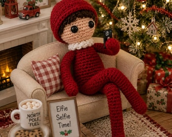 May include: A crocheted doll with brown hair and a red hat, wearing a red outfit with a white collar, sits on a small beige sofa holding a black smartphone. A sign in front reads "Elfie Selfie Time!" next to a mug labeled "NORTH POLE FUEL" with cookies.