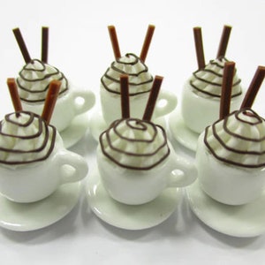 K&ouml;nnte beinhalten: Sechs wei&szlig;e, miniaturgro&szlig;e Keramikbecher mit brauner Schokoladensauce und Schlagsahne obenauf. Jeder Becher hat zwei braune St&auml;bchen, die aus der Schlagsahne herausragen. Die Becher stehen auf wei&szlig;en Untertassen.