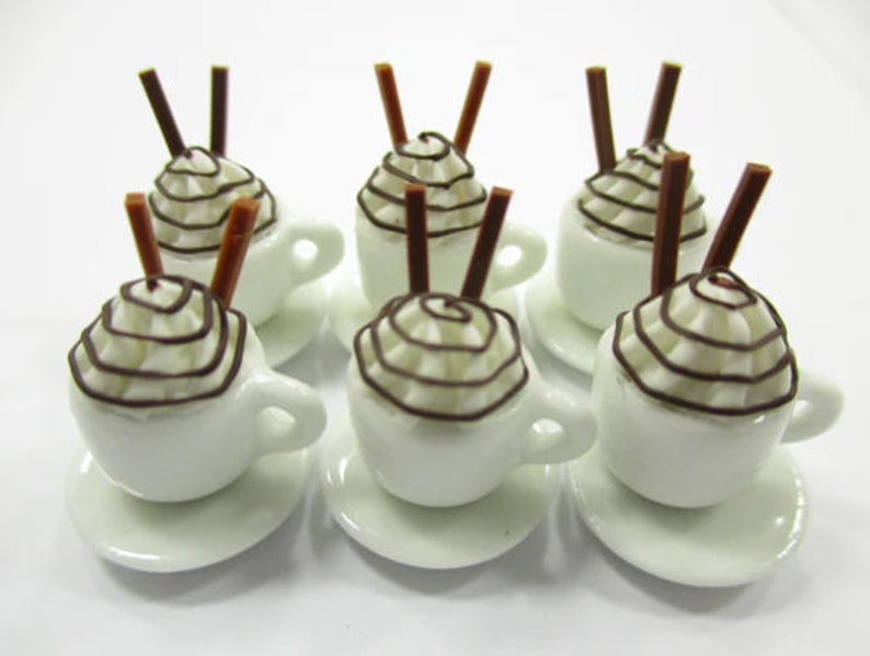 K&ouml;nnte beinhalten: Sechs wei&szlig;e, miniaturgro&szlig;e Keramikbecher mit brauner Schokoladensauce und Schlagsahne obenauf. Jeder Becher hat zwei braune St&auml;bchen, die aus der Schlagsahne herausragen. Die Becher stehen auf wei&szlig;en Untertassen.